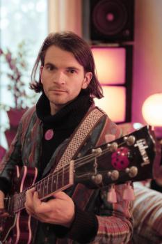 Preview: Bandfoto Harry-Hudson-Taylor: Musiker mit Gitarre im Innenraum (Porträt) Eine Person mit schulterlangem dunklem Haar sitzt in einem Innenraum und hält eine E-Gitarre. Sie blickt ruhig in die Kamera. Die Person trägt einen gemusterten Mantel und einen dunk